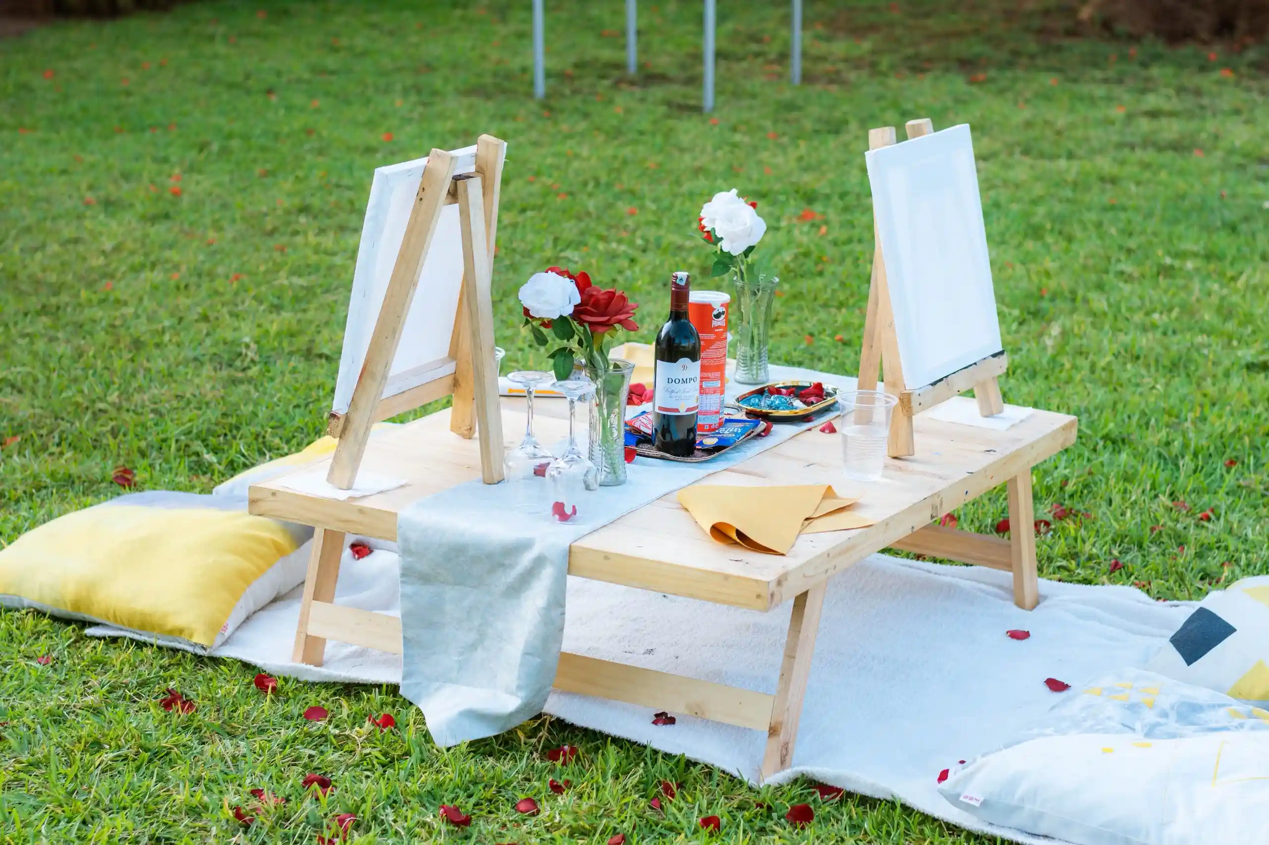 Two easels set for a couples painting date, with wine, roses and rose petals on the grass